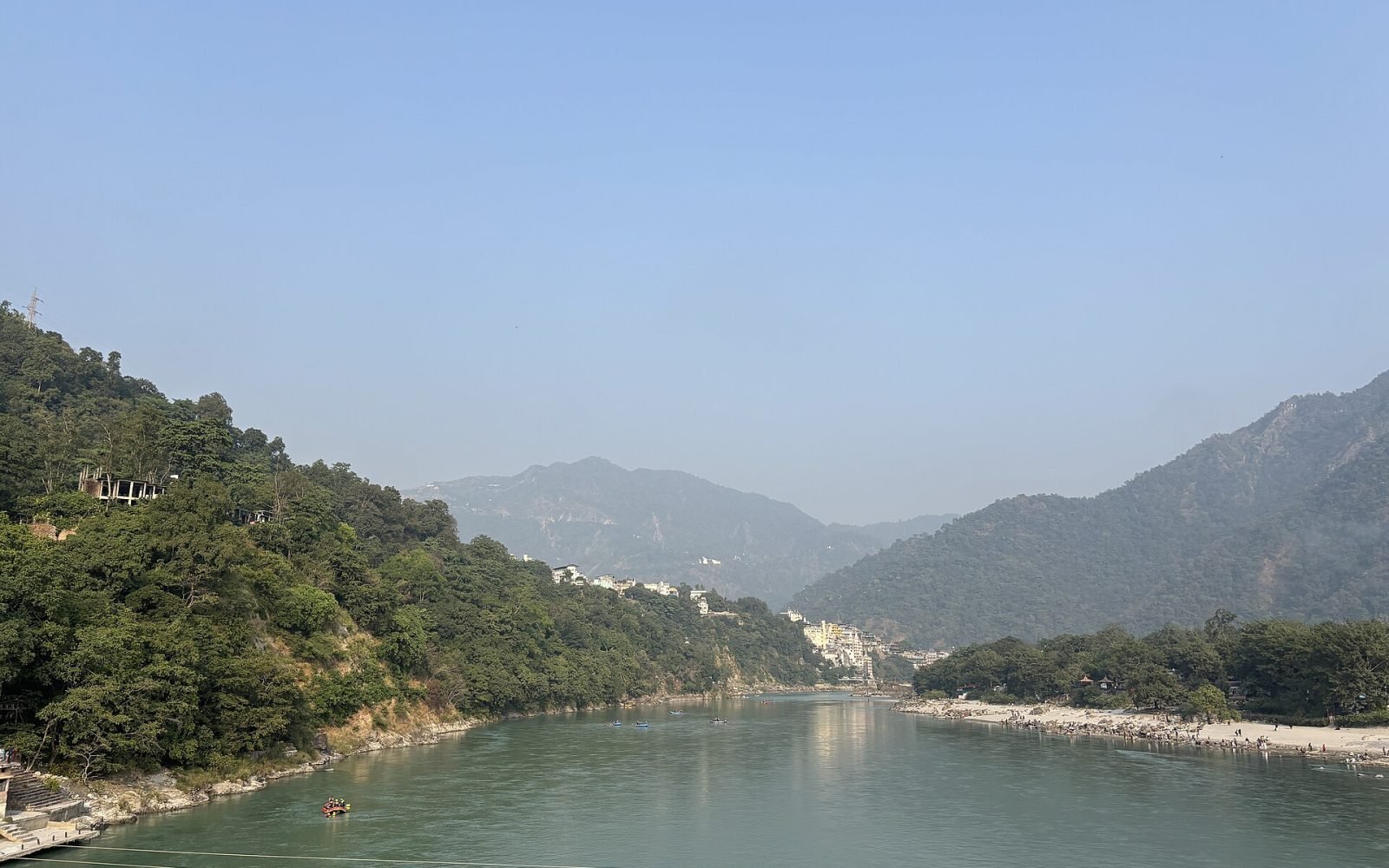 The sacred River Ganga flows between forested hills at Rishikesh, the water calm beneath the Himalayan foothills.